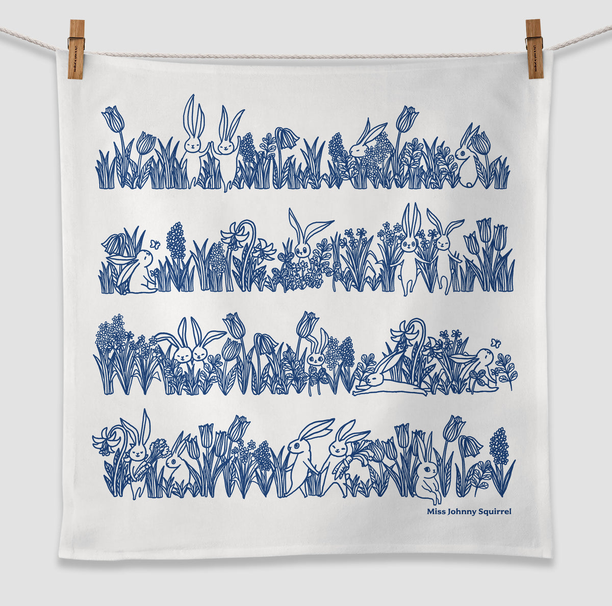 White 20x20 tea towel with blue line art illustration of bunnies sitting and hopping among tulips, grasses, and garden flowers, with Miss Johnny Squirrel logo.