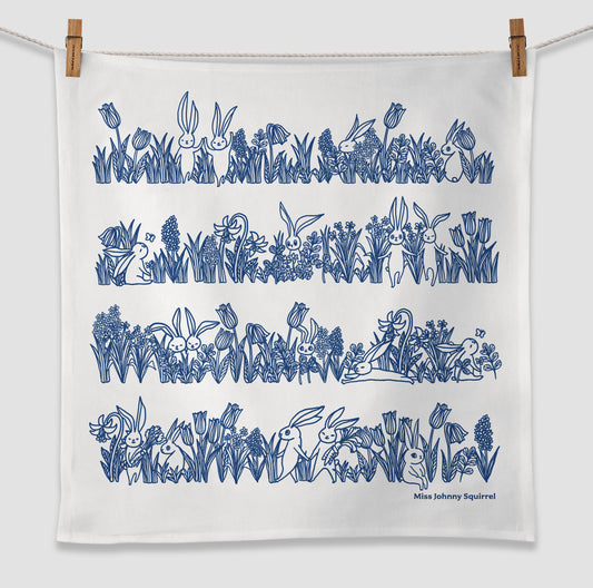 White 20x20 tea towel with blue line art illustration of bunnies sitting and hopping among tulips, grasses, and garden flowers, with Miss Johnny Squirrel logo.