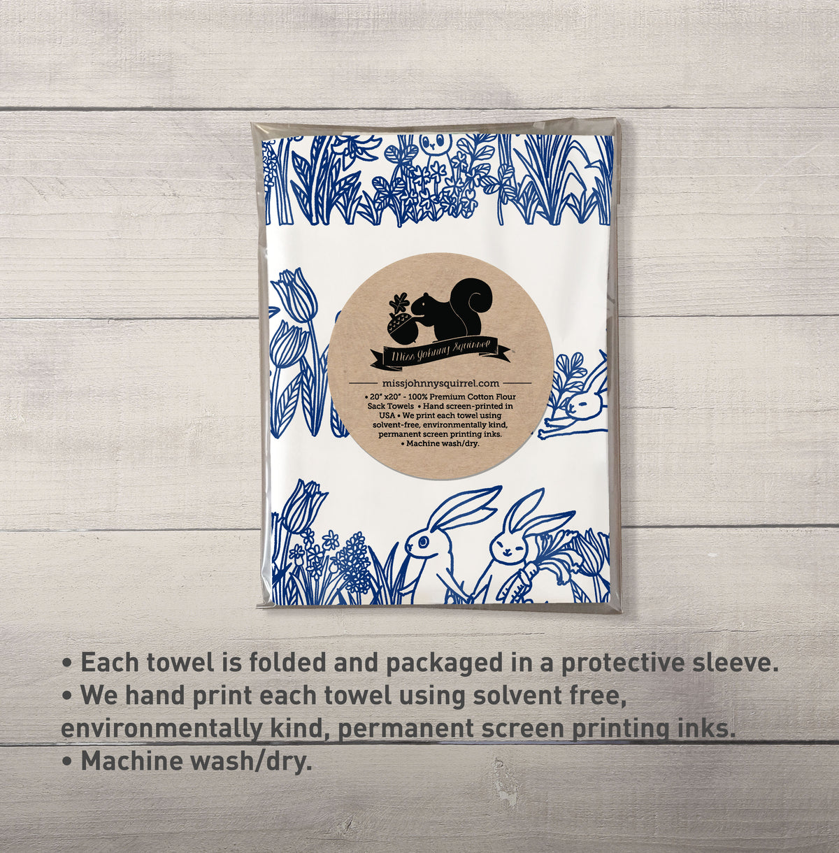 White 20x20 tea towel with blue line art illustration of bunnies sitting and hopping among tulips, grasses, and garden flowers, with Miss Johnny Squirrel logo.