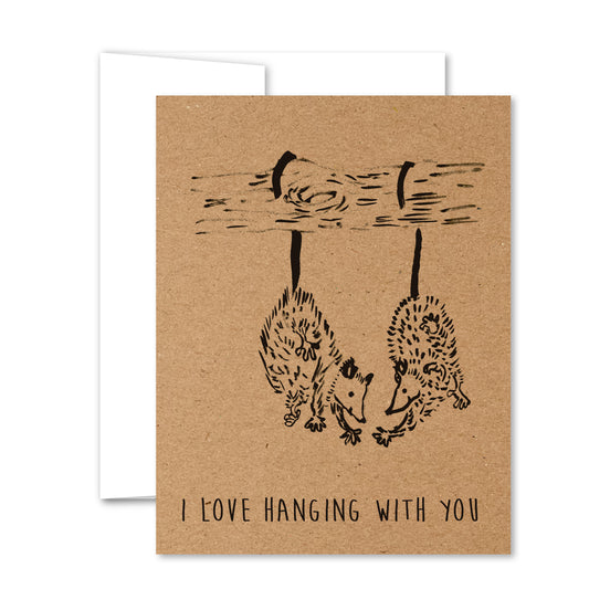 Kraft paper greeting card featuring two illustrated possums hanging by their tails from a branch, facing each other affectionately above the caption: “I love hanging with you.”