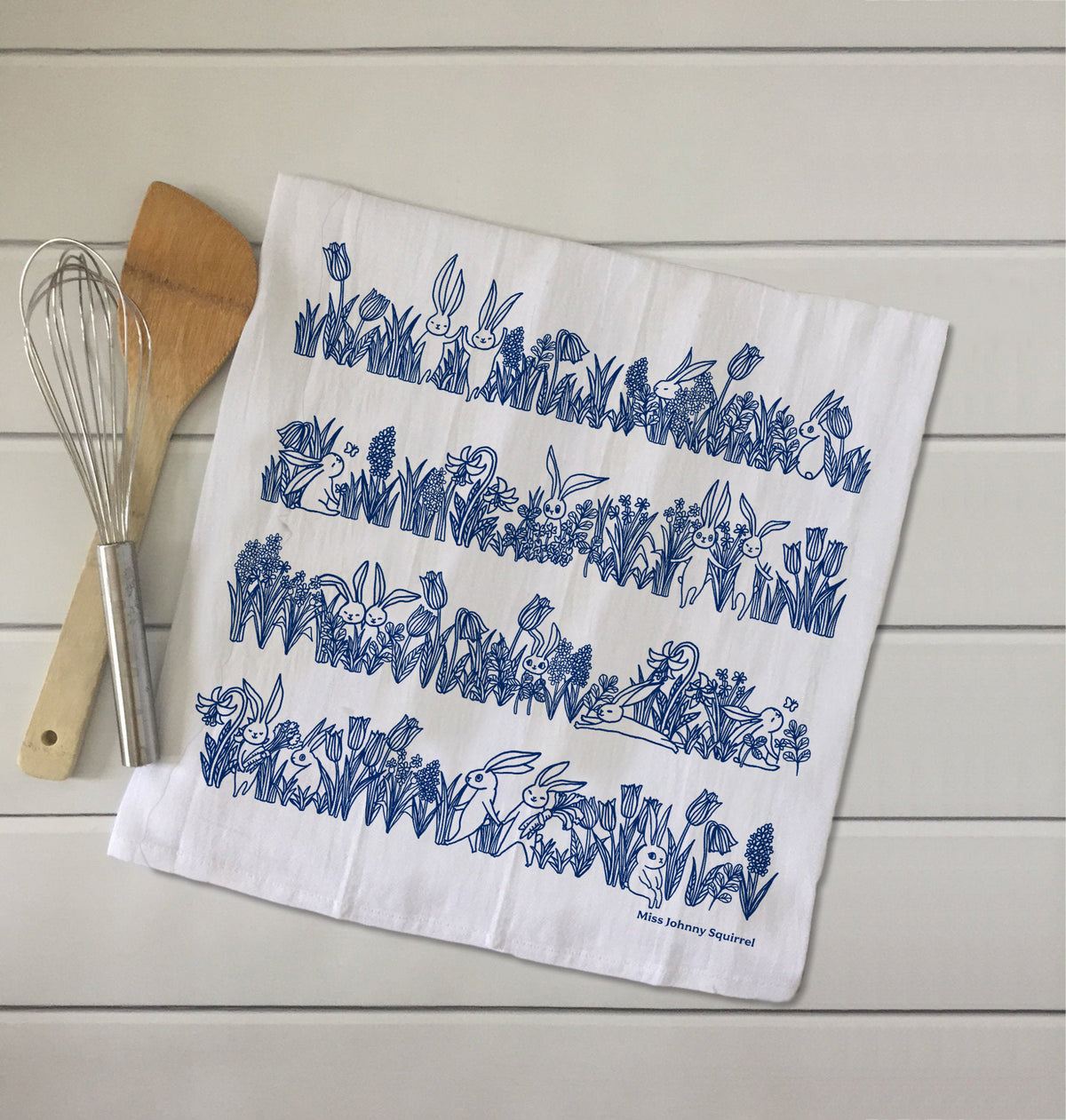 White 20x20 tea towel with blue line art illustration of bunnies sitting and hopping among tulips, grasses, and garden flowers, with Miss Johnny Squirrel logo.